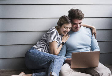Glückliches junges Paar sitzt vor einer Wand und benutzt einen Laptop - UUF28842