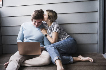 Smiling young woman kissing man using laptop on balcony - UUF28843