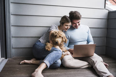 Lächelnder Mann und Frau, die einen Laptop benutzen und mit einem Hund vor einer Wand sitzen - UUF28845