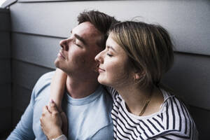 Lächelnde Frau und Mann sitzen mit geschlossenen Augen an einer Mauer auf einem Balkon - UUF28850