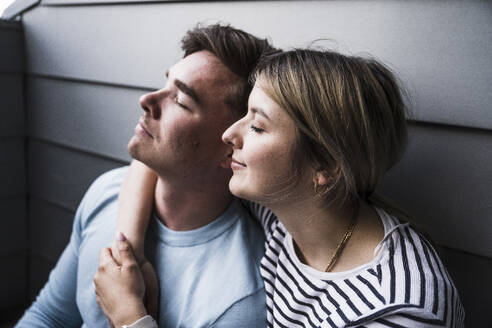 Smiling woman and man sitting with eyes closed by wall on balcony - UUF28850