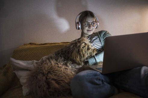 Smiling young woman wearing wireless headphones using laptop at home - UUF28853