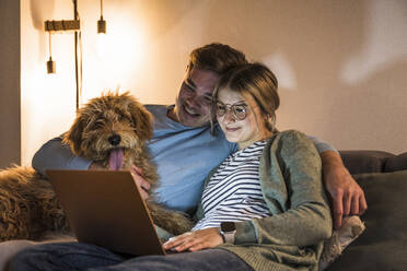 Lächelndes Paar, das einen Laptop benutzt und mit einem Hund auf dem Sofa zu Hause sitzt - UUF28856
