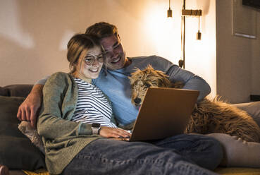 Lächelndes junges Paar, das einen Laptop benutzt und mit einem Hund auf dem Sofa zu Hause sitzt - UUF28857
