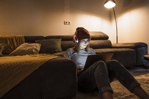 Young woman wearing wireless headphones and using laptop in living room - UUF28866