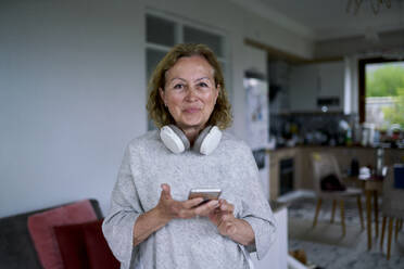 Smiling senior woman standing with smart phone at home - ANNF00289