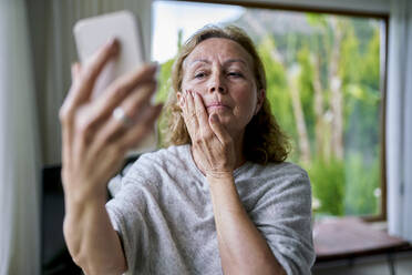 Ältere Frau schaut auf ihr Smartphone und berührt ihr Gesicht zu Hause - ANNF00296