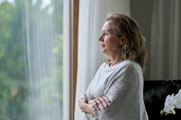 Thoughtful senior woman standing with arms crossed and looking through window at home - ANNF00298