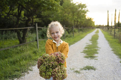 Smiling girl holding Hydrangea flower standing on footpath - SVKF01444