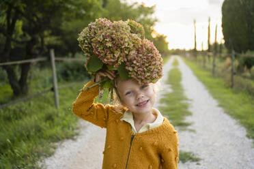Lächelndes Mädchen, das eine Hortensienblüte über den Kopf hält, auf einem Fußweg - SVKF01445