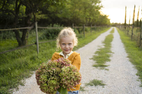 Smiling blond girl holding Hydrangea flower on footpath - SVKF01446