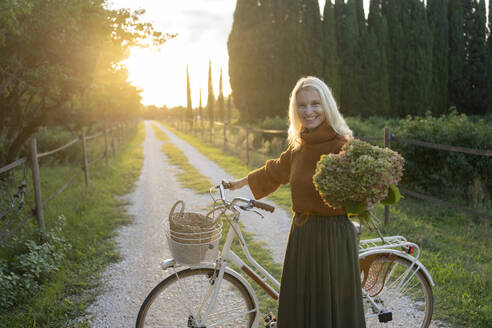 Happy woman with flower bunch standing next to bicycle on footpath - SVKF01449