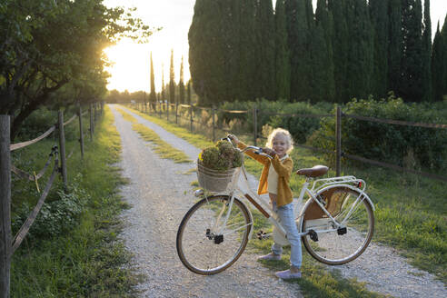 Smiling girl standing next to bicycle on footpath - SVKF01450