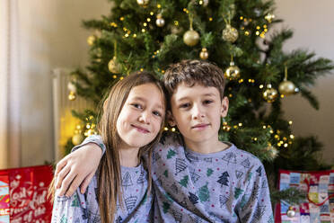 Lächelnde Schwester und lächelnder Bruder sitzen vor dem Weihnachtsbaum zu Hause - WPEF07418