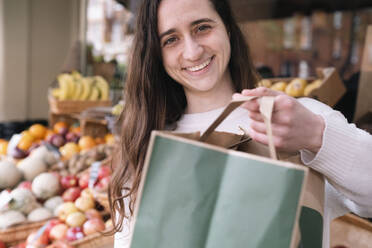 Happy young grocer standing with paper bag at vegetable stall - AMWF01400