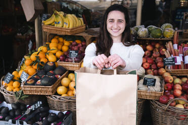 Happy young grocer standing with paper bag at vegetable shop - AMWF01402