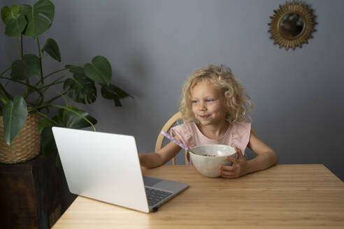 Smiling blond girl using laptop and having breakfast at dining table - SVKF01459