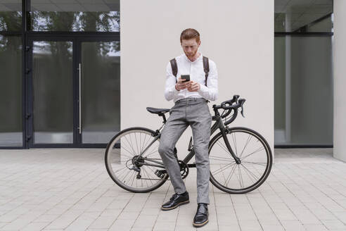 Geschäftsmann, der ein Smartphone benutzt und sich auf ein Fahrrad in der Nähe des Büros stützt - OSF01660