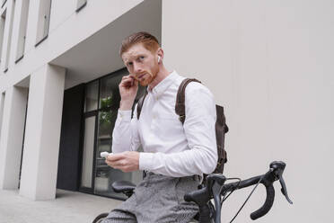 Geschäftsmann auf dem Fahrrad sitzend mit drahtlosen In-Ear-Kopfhörern in der Nähe des Büros - OSF01663