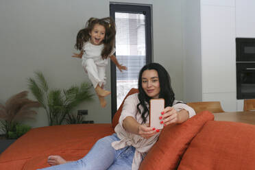 Eine Mutter fängt einen lustigen Moment ein, als ihre Tochter auf dem Sofa zu Hause herumspringt und ein Selfie macht - SYEF00426