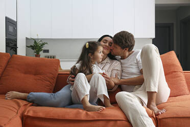 Man kissing woman sitting with daughter on sofa at home - SYEF00431