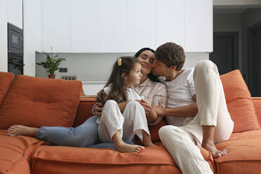 Mann küsst Frau, die mit ihrer Tochter zu Hause auf dem Sofa sitzt - SYEF00431