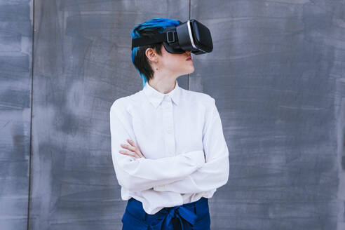 Businesswoman with arms crossed wearing virtual reality headset in front of wall - MGRF01020