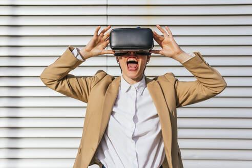 Businesswoman wearing virtual reality headset screaming in front of wall - MGRF01023