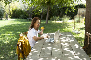 Smiling freelancer sitting with coffee cup and working on laptop - SVKF01465