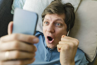 Excited man holding smart phone lying down on bed at home - TYF00793