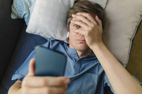 Worried man looking at smart phone lying on bed - TYF00794
