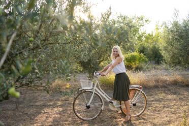 Happy blond woman with bicycle in olive farm - SVKF01468