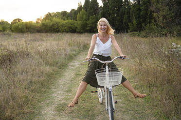 Happy mature woman riding bicycle on field - SVKF01469