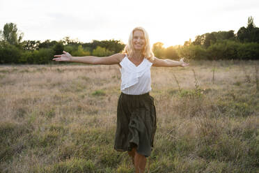 Glückliche Frau, die mit ausgestreckten Armen auf einem Feld bei Sonnenuntergang spazieren geht - SVKF01471