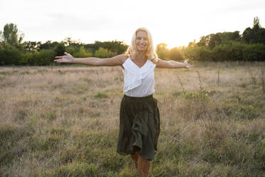 Happy woman walking with arms outstretched on field at sunset - SVKF01471