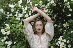 Junge Frau mit geschlossenen Augen und erhobenen Armen an einem weißen Blumenstrauch - SEAF01971