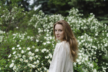 Young woman with long blond hair standing by white flower bush - SEAF01974