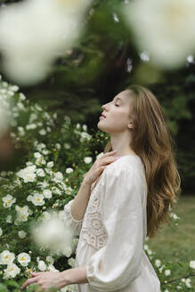 Junge Frau mit geschlossenen Augen an einem weißen Blumenstrauch stehend - SEAF01976