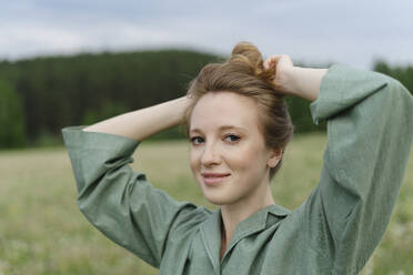 Lächelnde junge Frau beim Binden eines Haarknoten auf einem Feld - SEAF01983