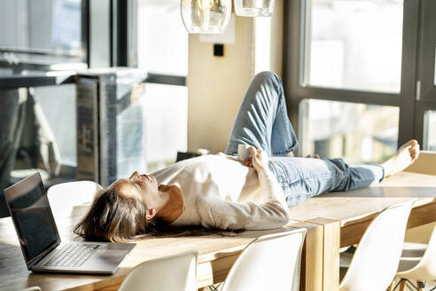 Businesswoman lying on table with coffee cup and laptop - PESF03973