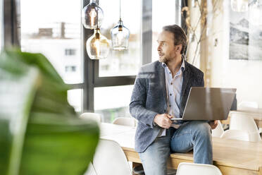 Businessman sitting with laptop on table in office cafeteria - PESF03978