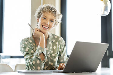 Glückliche blonde Geschäftsfrau sitzt mit Laptop am Tisch - PESF03992