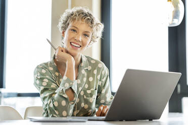 Happy blond businesswoman sitting with laptop at table - PESF03992