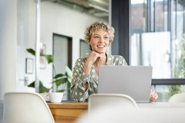 Thoughtful businesswoman sitting with laptop in office cafeteria - PESF03994