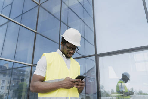 Ingenieur in reflektierender Kleidung, der mit seinem Smartphone vor einem Glasgebäude SMS schreibt - OSF01694