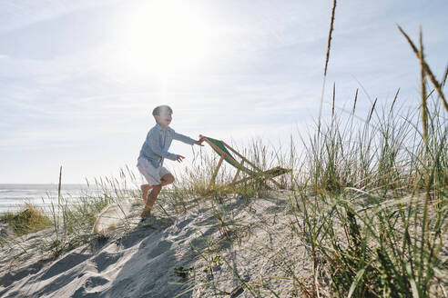 Playful boy by folding chair on sandy beach - ASGF03746