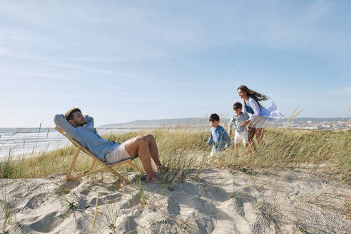 Vater entspannt sich auf einem Stuhl mit seiner Familie, die am Strand spielt, an einem sonnigen Tag - ASGF03753