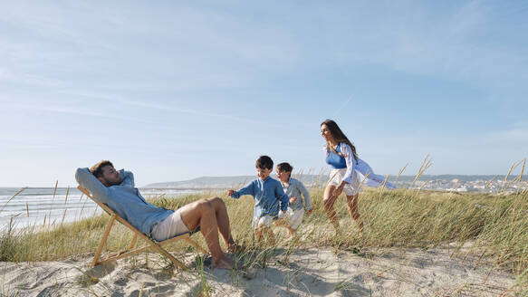 Familie läuft auf den Vater zu, der sich an einem sonnigen Tag am Strand auf einem Stuhl ausruht - ASGF03754