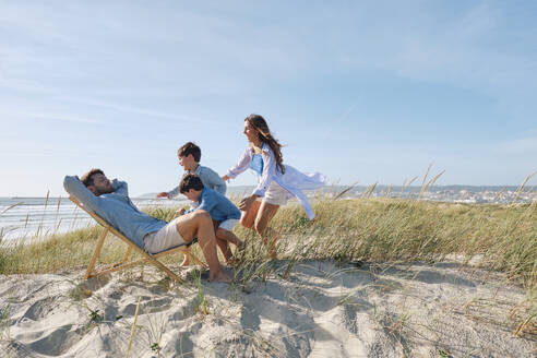 Family running towards father relaxing on folding chair at beach - ASGF03755