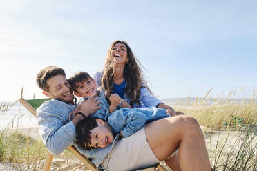Glückliche Familie umarmt Vater sitzt auf Stuhl am Strand - ASGF03756